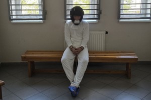 na zdjęciu widzimy mężczyznę podejrzanego o dokonanie zabójstwa który siedzi  na ławce w pomieszczeniu dla osób zatrzymanych. Mężczyzna ma założone kajdanki zespolone na ręce oraz nogi.