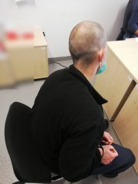 Na zdjęciu widać zatrzymanego mężczyznę, który siedzi na krześle z boku biurka. Mężczyzna ma założone kajdanki, kajdanki założone są na ręce trzymane z przodu. Policjant pionu kryminalnego przesłuchuje mężczyznę siedząc za biurkiem.