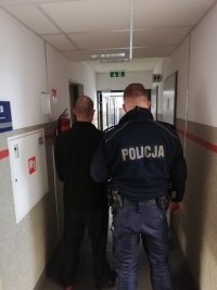 Korytarzem Komendy Miejskiej Policji w Dąbrowie Górniczej umundurowany policjant prowadzi zatrzymanego.