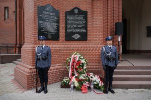 Policjanci stojący pod tablicą pamiątkową gdzie złożono wieńce