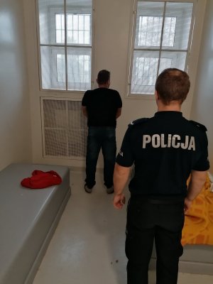 Umundurowany policjant w pomieszczeniu dla osób zatrzymanych wraz z zatrzymanym  mężczyzną, obaj stojący tyłem.