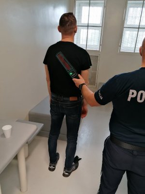 Mężczyzna w policyjnym areszcie, stojący tyłem. Umundurowany policjant sprawdza urzadzeniem czy w/w nie posiada przy sobie metalowych przedmiotów.