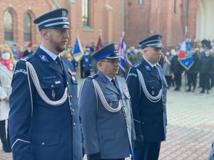 Zdjęcia przedstawiające trzech umundurowanych galowo policjantów stojących na baczność, w tle poczty sztandarowe.