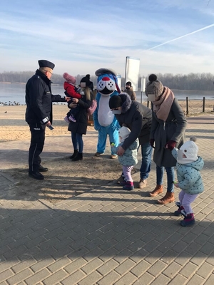 Umundurowany policjant oraz maskotka policyjna Sznupek nad zbiornikiem wodnym Pogoria III, podczas spotkania z osobami morsującymi, oddajacymi krew oraz w trakcie wreczania odblasków turystom wypoczywającym zimą nad wodą.