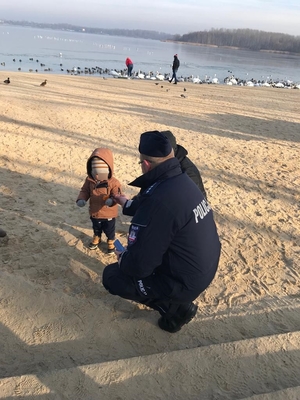 Umundurowany policjant oraz maskotka policyjna Sznupek nad zbiornikiem wodnym Pogoria III, podczas spotkania z osobami morsującymi, oddajacymi krew oraz w trakcie wreczania odblasków turystom wypoczywającym zimą nad wodą.