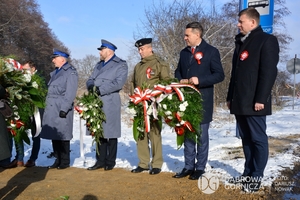 Na zdjęciu widzimy kilka osób składających hołd poległym przy krzyżu, wśród nich 2 umundurowanych policjantów i 1 umundurowany wojskowy.