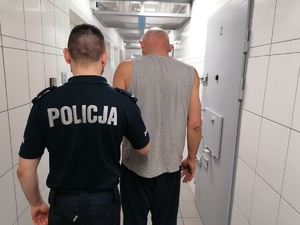 Stojący tyłem umundurowany policjant wraz z zatrzymanym mężczyzną w korytarzu pomieszczenia dla osób zatrzymanych.