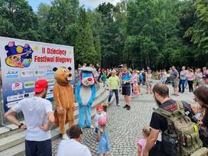 Umundurowani Policjanci i maskotka policyjna Sznupek , radiowóz oznakowany i motocykl służbowy w trakcie trwania 2 Dziecięcego Festiwalu Biegowego.