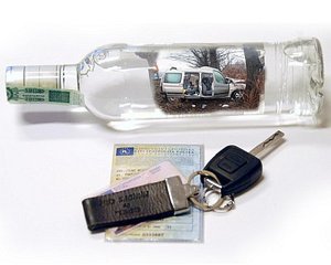 Na zdjęciu widać butelkę szklaną w tle której jest rozbity pojazd. pod tym obrazkiem jest kluczyk do samochody przykrywający prawo jazdy i dowód rejestracyjny.