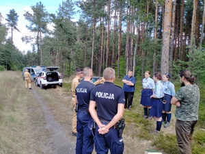 Umundurowani policjanci , strażacy i harcerze podczas sprawdzania obozowiska usytuowanego w lesie.