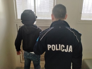 Umundurowany policjant stojący z zatrzymanym w celi.  Na głowie zatrzymanego kask bezpieczeństwa.