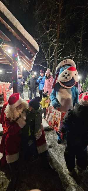 Umundurowani Policjanci i Sznupek wraz ze Świętym Mikołajem wręczają prezenty dzieciom.