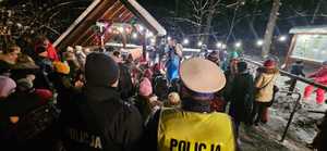 Umundurowani Policjanci stojący tyłem przyglądają się jak Sznupek wraz ze Świętym Mikołajem wręczają prezenty dzieciom.