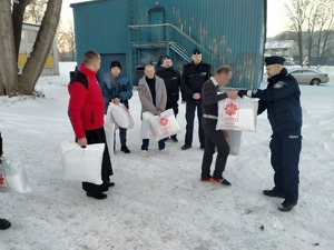 Umundurowani policjanci i ksiądz wraz z pracownikami Caritas wręczają paczkę bezdomnym.