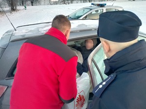 Umundurowany policjant i ksiądz wręczają paczkę bezdomnemu siedzącemu w samochodzie.