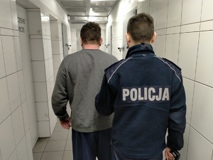 Mężczyzna prowadzony przez umundurowanego policjanta na korytarz pomieszczenia dla osób zatrzymanych.