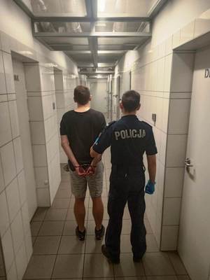 Na zdjęciu widzimy stojących tyłem - umundurowanego policjanta z zatrzymanym w korytarzu policyjnego aresztu.