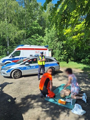 Na zdjęciu widać ratownika medycznego który udziela instruktarzu z pierwszej pomocy kobiecie. W tle widać umundurowanego policjanta oraz radiowóz policyjny i karetkę pogotowia.