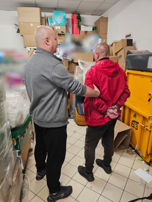 Nieumundurowany policjant stojący tyłem z mężczyzną który ma kajdanki na rękach trzymanych z tyłu.