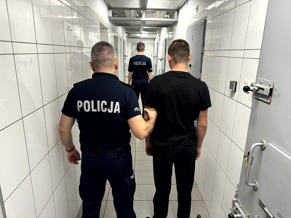 Dwóch umundurowanych policjantów prowadzących korytarzem zatrzymanego.