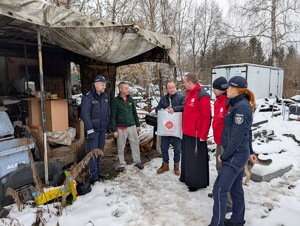 Umundurowani policjanci i pracownicy Caritas wraz z księdzem podczas rozdawani kocy, śpiworów dla bezdomnych.