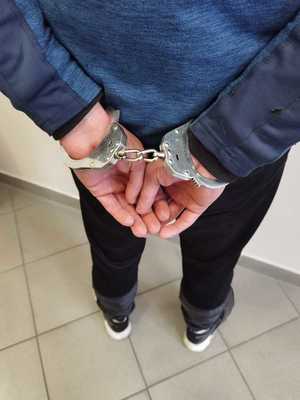 Na zdjęciu widzimy mężczyznę stojącego tyłem. Mężczyzna ma założone kajdanki na ręce trzymane z tyłu.