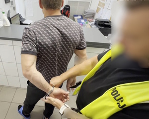 Zatrzymany stojący tyłem. Nieumundurowany policjant w odblaskowej kamizelce zakłada mężczyźnie kajdanki na ręce trzymane z tyłu.