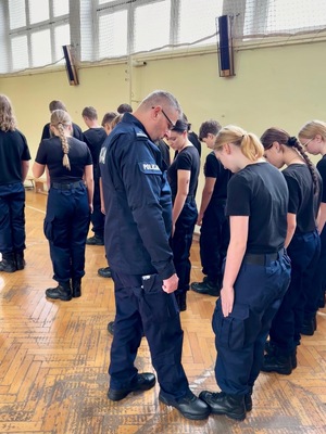Umundurowani policjanci i kadeci na zajęciach z musztry policyjnej w sali gimnastycznej.