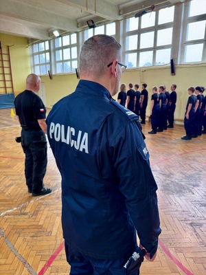 Umundurowani policjanci i kadeci na zajęciach z musztry policyjnej w sali gimnastycznej.