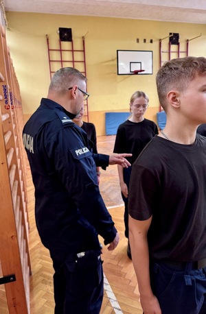 Umundurowani policjanci i kadeci na zajęciach z musztry policyjnej w sali gimnastycznej.