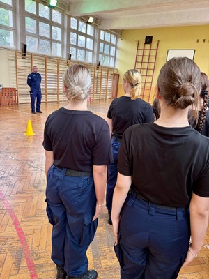 Umundurowani policjanci i kadeci na zajęciach z musztry policyjnej w sali gimnastycznej.
