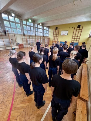 Umundurowani policjanci i kadeci na zajęciach z musztry policyjnej w sali gimnastycznej.