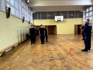 Umundurowani policjanci i kadeci na zajęciach z musztry policyjnej w sali gimnastycznej.