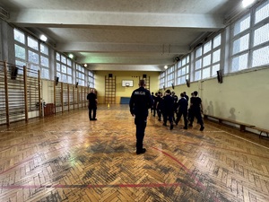 Umundurowani policjanci i kadeci na zajęciach z musztry policyjnej w sali gimnastycznej.