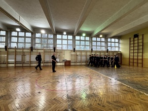 Umundurowani policjanci i kadeci na zajęciach z musztry policyjnej w sali gimnastycznej.