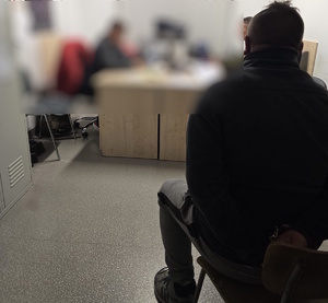 Mężczyzna siedzący tyłem do biurka przesłuchiwany przez nieumundurowanych policjantów.