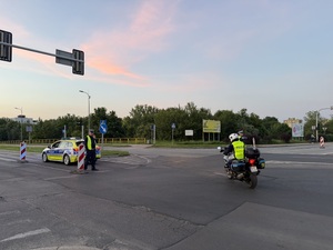 Zdjęcie przedstawia jadący motocykl policyjny, radiowóz policyjny wraz z umundurowanymi policjantami podczas zabezpieczenia I Maratonu Zagłębiowskiego w dniu 23 maja 2026 roku.