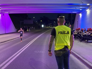 Zdjęcie przedstawia stojącego tyłem policjanta w kamizelce odblaskowej wraz z biegnącym uczestnikiem biegu podczas zabezpieczenia I Maratonu Zagłębiowskiego w dniu 23 maja 2026 roku.