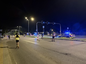 Zdjęcie przedstawia biegnących uczestników wraz z radiowozami policyjnymi w tle podczas zabezpieczenia I Maratonu Zagłębiowskiego w dniu 23 maja 2026 roku.