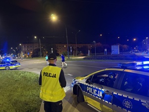 Zdjęcie przedstawia radiowóz policyjny wraz z umundurowanym policjantem i biegnącymi uczestnikami biegu w tle podczas zabezpieczenia I Maratonu Zagłębiowskiego w dniu 23 maja 2026 roku.