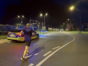 Zdjęcie przedstawia biegnącego uczestnika biegu wraz z częścią radiowozu policyjnego podczas zabezpieczenia I Maratonu Zagłębiowskiego w dniu 23 maja 2026 roku.