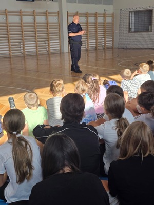 Umundurowany policjant na spotkaniu z dziećmi w szkole podstawowej - sala gimnastyczna wykład.